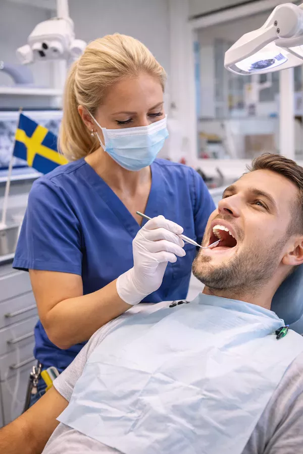 Tandvårdsklinik i Sverige med patient i behandlingsstol – undersökning av tandläkare i blå vårdkläder och svensk flagga i bakgrunden.
